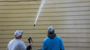 Two young men standing by the side of a yellow house. One is power washing the siding with a long nozzle.