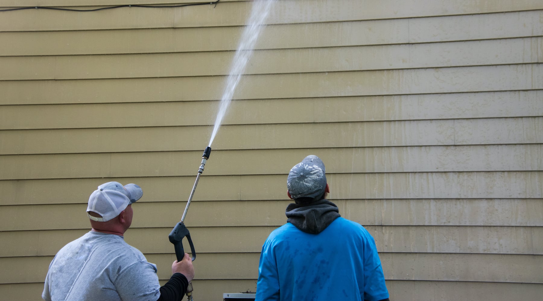 Two young men standing by the side of a yellow house. One is power washing the siding with a long nozzle.