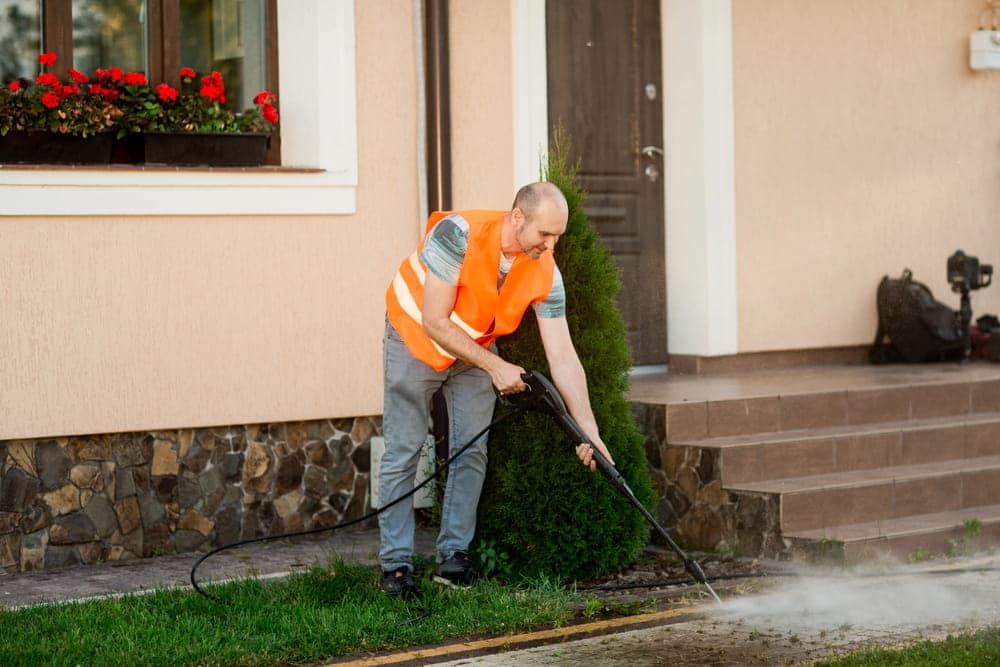 A man in an orange vest cleans a tile of grass in his yard near the house